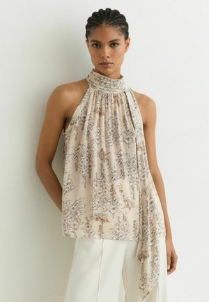 Femeie purtând un top bej fără mâneci cu imprimeu floral și pantaloni albi, stând în fața unui perete alb simplu.