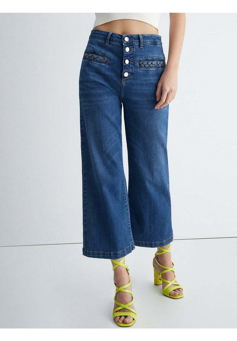 Jeans niebieskie o wysokim stanie i szerokich nogawkach z przednim zapięciem na guziki oraz plecionymi detalami kieszeni, noszone z neonowozielonymi sandałami na obcasie z paskami.