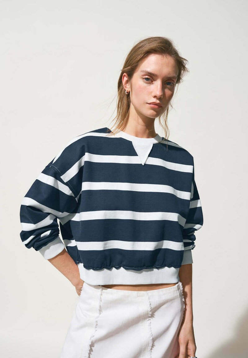 Jeune femme aux cheveux clairs portant un sweat court rayé bleu marine et blanc et une jupe blanche, debout avec une main sur la hanche.