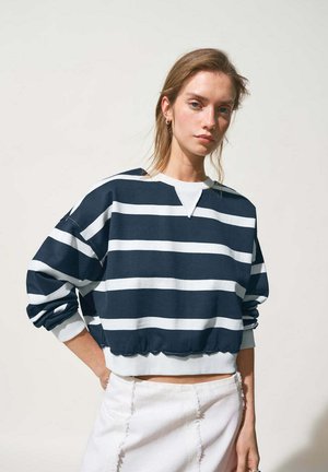 Giovane donna con capelli chiari che indossa una felpa corta a righe blu navy e bianche e una gonna bianca, in piedi con una mano sui fianchi.