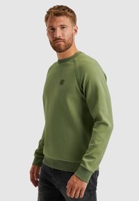 Groene sweatshirt met een geribbelde ronde hals en lange mouwen, voorzien van een subtiele logo op de borst en gebreide manchetten, weergegeven met donkere jeans.