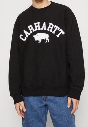 Person trägt schwarzen Sweatshirt mit weißem "CARHARTT" Schriftzug und Bison-Logo, kombiniert mit blauen Jeans, vor einfarbigem Hintergrund.