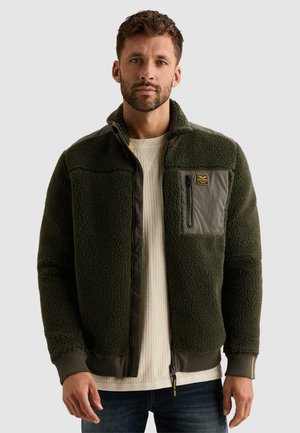 Homme portant une veste polaire vert foncé avec une poche poitrine zippée par-dessus un pull en maille crème, debout devant un fond clair uni.