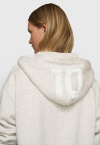 Sudadera gris claro con una textura suave, que presenta un "10" blanco de gran tamaño en la parte trasera de la capucha. Diseño sencillo y ajuste casual.