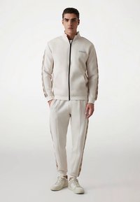 Kremowa bluza dresowa na zamek z detalami w postaci taśm i logo, wykonana z gładkiego materiału. Dopasowane spodnie z bocznymi paskami.