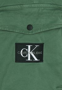 Tissu vert avec une poche ornée d'un patch noir affichant "CALVIN KLEIN JEANS" et un logo "cK" blanc. Détails de couture visibles.