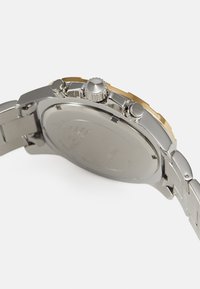 Silverfärgad klocka i rostfritt stål med en guld-accentuerad bezel. Har en slät, polerad baksida och tre framträdande tryckknappar.