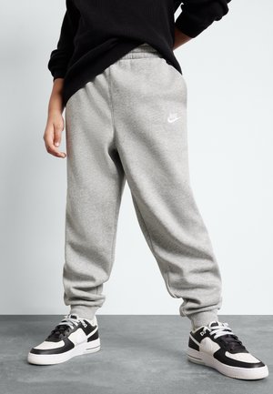 Osoba ubrana w szare spodnie dresowe Nike i czarno-białe sneakersy Nike, stojąca na szarej podłodze na tle jednolitego jasnego tła.