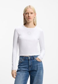 Ξανθιά γυναίκα που φοράει ένα εφαρμοστό λευκό crop top με μακριά μανίκια και μικρό κυκλικό σχέδιο, καθώς και ψηλόμεσο μπλε τζιν, με το ένα χέρι στην τσέπη.