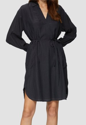 Vrouw draagt een zwarte jurk tot op de knie met lange mouwen en subtiele strepen, V-hals en een strikceintuur in de taille, staat met handen in de zij.