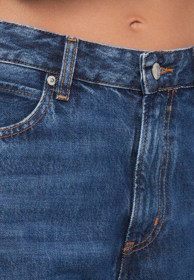 Prim-plan al secțiunii medii îmbrăcată în blugi albaștri din denim, cu nasture și fermoar, arătând pielea expusă deasupra taliei și buricul.