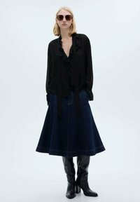 Crna svila bluza s detaljem volana i vezanjem, uparen s tamnoplavom A-linijom suknjom od trapera. Model nosi crne čizmice do koljena i oversized naočale.