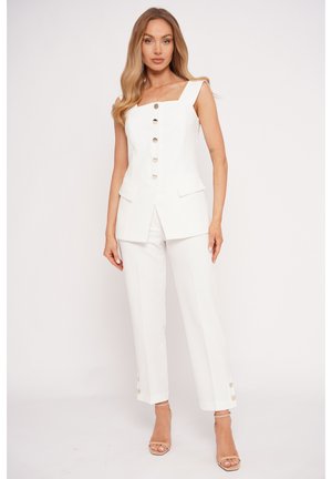 Kobieta w białej, bezrękawnikowej koszuli na guziki i dopasowanych krótkich spodniach, nosząca beżowe sandały na obcasie, stoi na jednolitym jasnym tle.