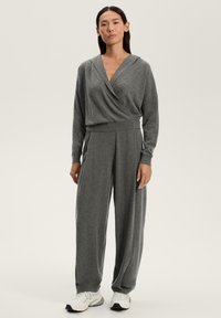 Grå, mjuk tygdräkt med djup V-ringning och huva. Löst sittande med lång ärm och vida byxor. Bärs med vita sportskor.