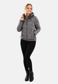 Woman smiling, wearing a gray puffer jacket with hood, black pants, and black ankle boots, standing against a plain light background.
