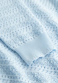 Lyseblå strikkegenser med intrikate blondermønstre og ribbet mansjett, som viser detaljert tekstur og håndverk.