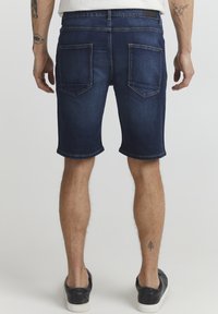 Tumši zili denim šorti ar salocītu apmalīti, ar divām aizmugures kabatām un minimālu šuvju detaļu, apvienoti ar melniem sporta apaviem.