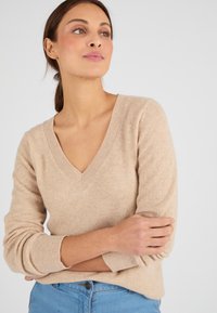 Maglione beige a V con maniche lunghe in morbido tessuto texturizzato, con polsini a costine e una vestibilità rilassata. Abbinato a jeans azzurri.