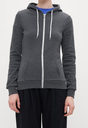 Person trägt einen dunkelgrauen Kapuzenpullover mit Reißverschluss, weißen Kordeln und Vordertaschen über einem blauen Hemd sowie schwarze Hose.