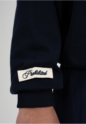 Ärmel eines dunkelmarineblauen Kleidungsstücks mit umgeschlagenem Bündchen, das ein weißes rechteckiges Stoffetikett zeigt, auf dem "Prohibited" in schwarzer Schrift aufgenäht ist.