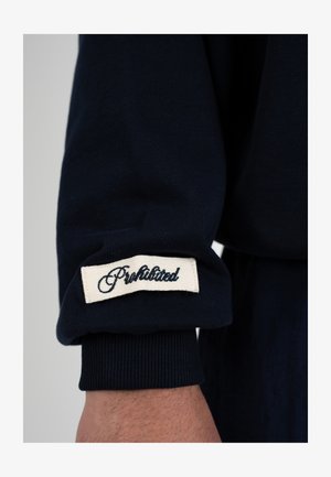 Mouw van een donkerblauwe kledingstuk met omgeslagen boord met een wit rechthoekig stoffen label waarop "Prohibited" met zwarte letters is gestikt.