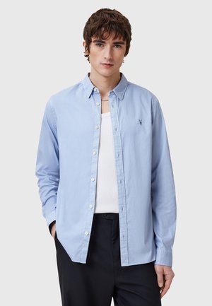 Camicia azzurro chiaro con bottoni, realizzata in cotone, dotata di logo sul petto, maniche lunghe e una vestibilità comoda. Abbinata a una canotta bianca sotto.