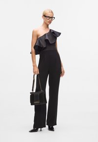 Svart jumpsuit med volang i satin på ena axeln, slim fit och lång benlängd. Matchad med en svart handväska och lackade klackar.