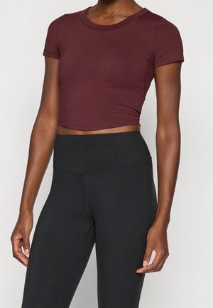 Vrouw draagt een aansluitende korte mouwen bordeauxrode crop top en hooggetailleerde zwarte leggings, staand tegen een effen lichte achtergrond.