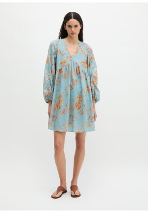 Vrouw staat en draagt een losse lichtblauwe jurk met oranje bloemmotief, lange pofmouwen en bruine platte sandalen tegen een witte achtergrond.
