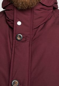 Giacca a piumino bordeaux con collo alto, caratterizzata da un bottone a pressione argento e un bottone marrone con iscrizione "HILFIGER". Texture liscia.