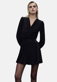 Abito nero a forfait con maniche lunghe, gonna plissettata e cintura con fibbia argentata. Tessuto liscio con una leggera lucentezza.