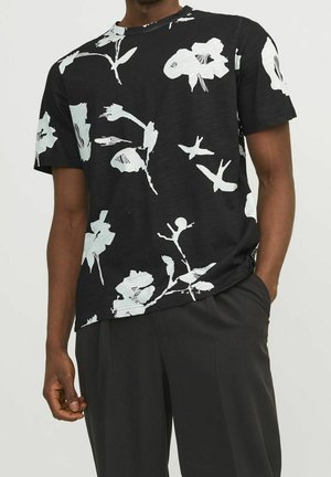 Homme portant une chemise noire à manches courtes avec de grandes impressions florales blanches, associée à un pantalon noir, debout avec une main dans la poche.