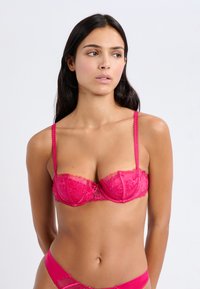 Soutien-gorge demi-cup en dentelle de couleur rose vif avec des motifs floraux, des bords festonnés et des bretelles délicates et ajustables. Texture de tissu douce.