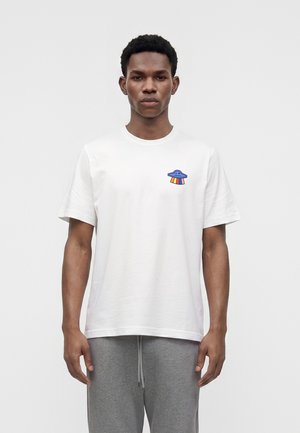 Homme portant un t-shirt blanc avec un petit design d’OVNI bleu et des rayons arc-en-ciel sur la poitrine, debout devant un fond gris clair uni.