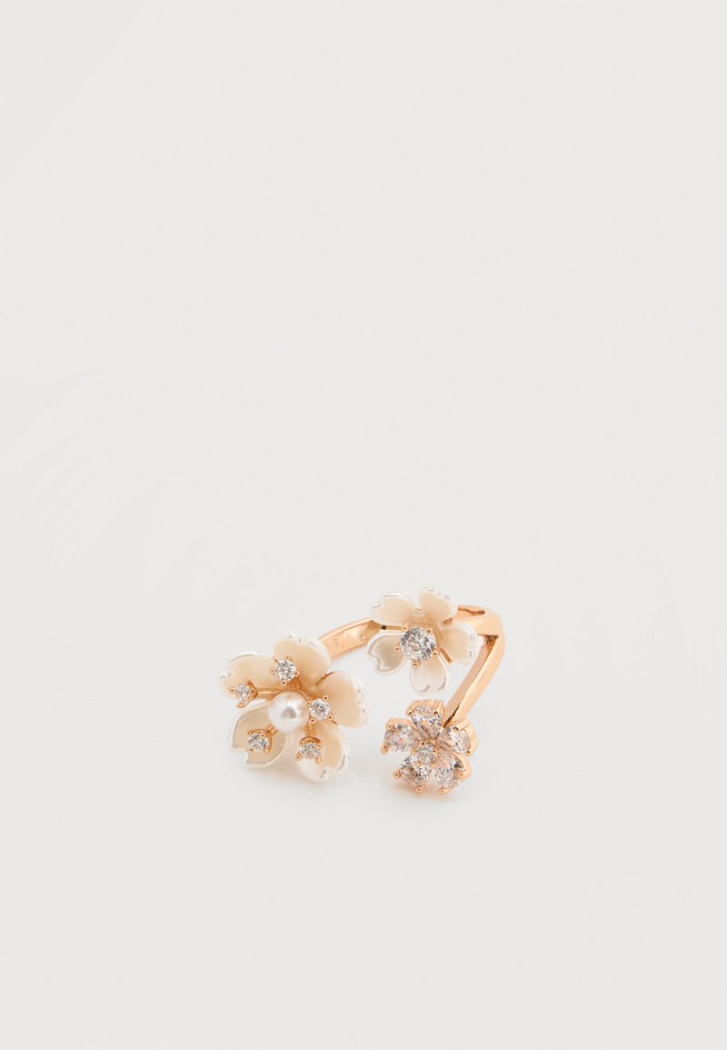 Bague florale en ton or avec trois fleurs, deux avec des centres en cristal et une avec un centre en perle, sur fond blanc.