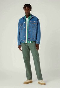 Μπλε τζιν μπουφάν με δύο τσέπες στο στήθος, πράσινο και λευκό ριγέ πουκάμισο από κάτω, ελαιόχρωμο παντελόνι και κρεμ αθλητικά παπούτσια.