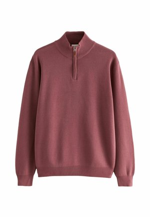 Homme portant un pull rose à fermeture éclair quart, un t-shirt blanc, un pantalon noir et des chaussures en cuir noires, debout avec une main dans la poche.