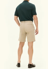 Pantaloni corti beige con due tasche posteriori, abbinati a una maglietta polo verde scuro. Il tessuto sembra liscio e i pantaloni corti hanno una vestibilità su misura.