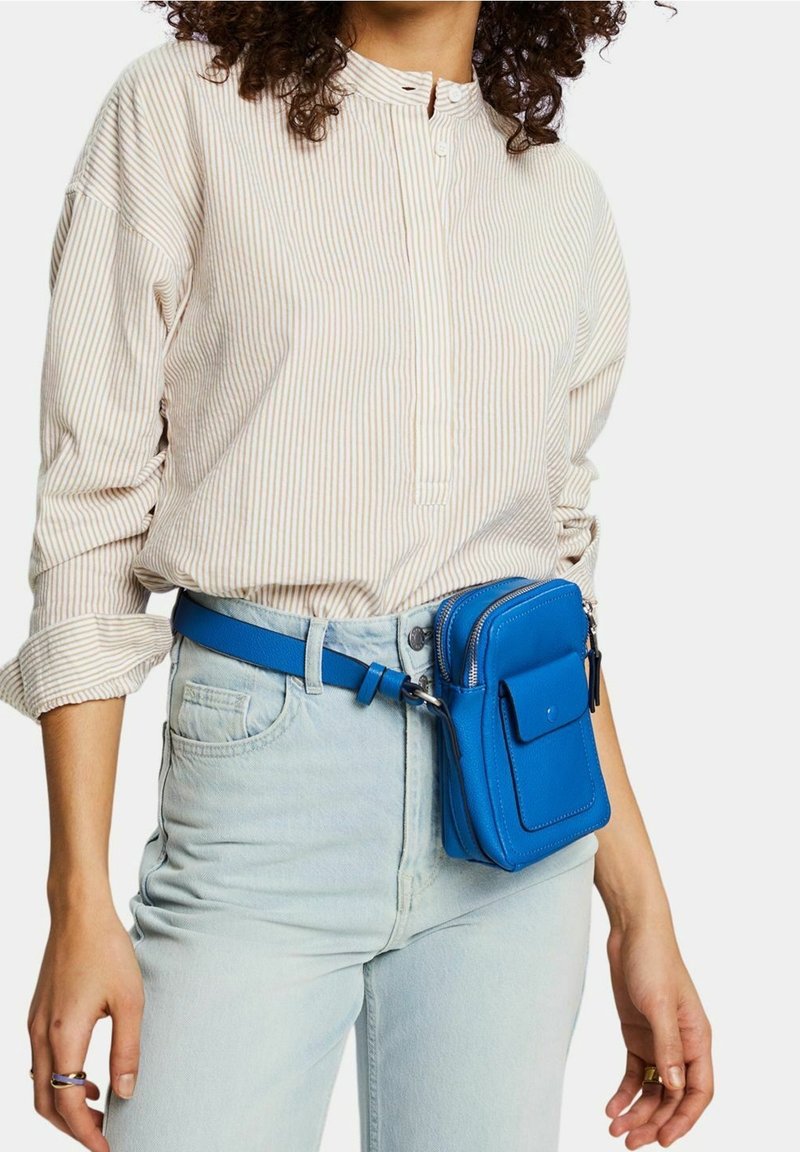Femme portant une chemise à manches longues à rayures claires et un jean bleu clair avec une ceinture-sac en cuir bleu vif à la taille.