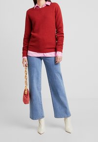 Maglione rosso con texture sopra una camicia rosa con colletto, abbinato a jeans a gamba larga blu, décolleté bianchi a punta e una borsa rossa con catena dorata.