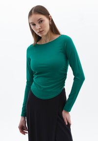 Maglietta a maniche lunghe con collo rotondo in tessuto verde uniforme, con dettagli arricciati sui lati, abbinata a una gonna nera. Tessuto morbido.