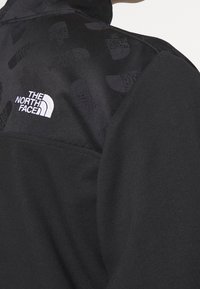 Giacca in pile nera con un pannello testurizzato caratterizzato da un motivo logo ripetuto sulla spalla, completa di un logo bianco sul petto.