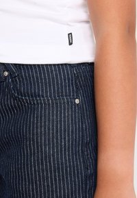 Lähikuva henkilöstä, joka on pukeutunut tummansinisiin pinstripe-farkkuihin ja valkoiseen paitaan, jossa on pieni "AMERICA"-lappu. Oikealla puolella näkyy kyynärvarsi.