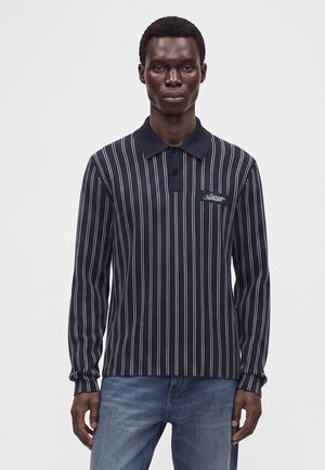 Homme portant un polo bleu marine à manches longues avec des rayures verticales blanches et un jean bleu clair, debout devant un fond blanc uni.
