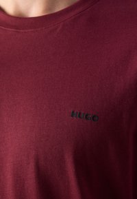 Close-up van een bordeauxrode crewneck shirt met een klein zwart "HUGO" tekstlogo op de linkerborst.