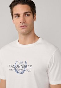 Junger Mann mit kurzen dunklen Haaren, der ein weißes T-Shirt mit dem Text "FACONNABLE UNIVERSITY CAMPUS 1950" und einer blauen Lorbeer-Grafik trägt.