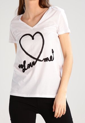 Kvinde iført en hvid V-hals t-shirt med et sort hjerte og teksten "Love me", kombineret med sorte bukser, stående mod en ensfarvet baggrund.