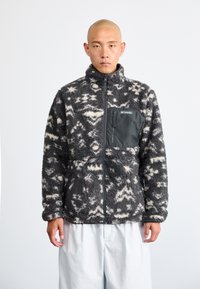 Fleecejacke mit einem schwarz-weißen abstrakten Muster, hohem Kragen, Reißverschlusstasche auf der Brust und langen Ärmeln. Weiches, strukturiertes Material.