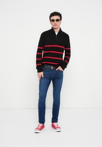 Μαύρο πουλόβερ με κόκκινες ρίγες, ψηλός γιακάς, κλείσιμο με φερμουάρ· μπλε τζιν, μαύρη ζώνη με μεταλλικά χτυπητά, κόκκινα αθλητικά παπούτσια, γυαλιά ηλίου.