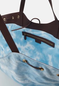 Sac fourre-tout ouvert avec poignées et garniture en cuir brun, présentant un extérieur en tissu tie-dye bleu et une poche intérieure zippée avec fermeture éclair dorée.